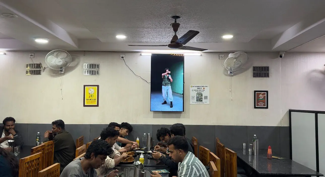 Diners at long tables beneath an AdVerse screen at Kannur Food Kitchen, Taverekere