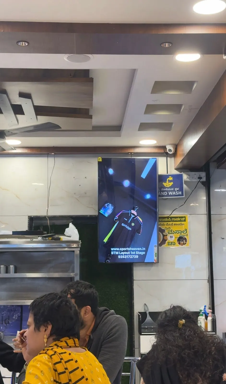 A portrait AdVerse screen above the counter at New Udupi Garden, Jayanagar, with diners in the foreground